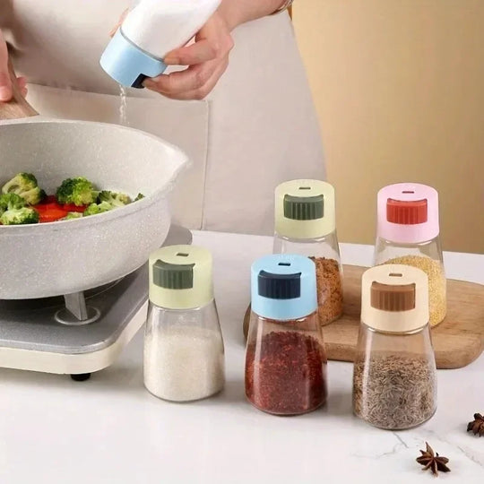 Person seasoning a dish with a salt shaker, surrounded by small spice bottles on a kitchen counter.