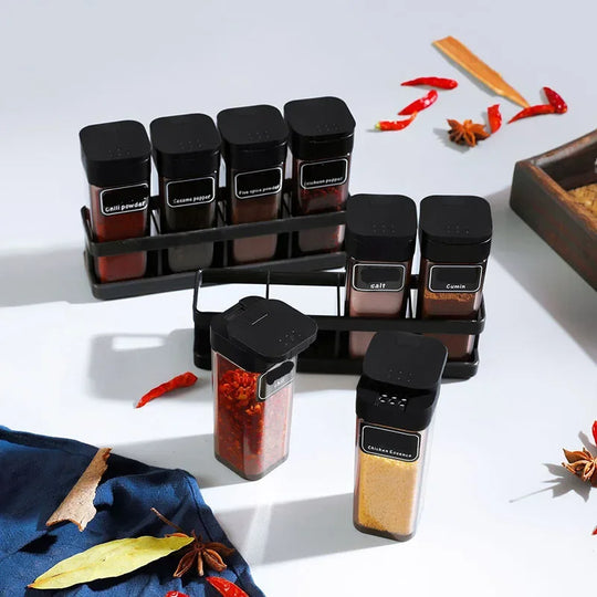 Set of spice jars with black lids on a white surface, surrounded by dried herbs and spices.