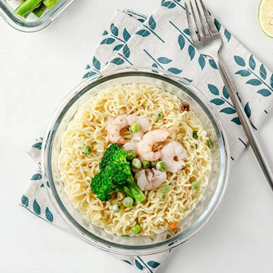 Glass bowl with noodles, shrimp, and broccoli on a white surface with a leaf-patterned napkin.
